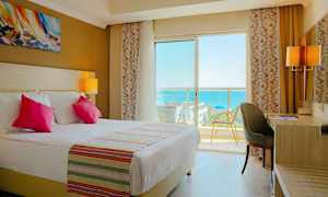 Hotellrum med dubbelsäng, skrivbord och balkongutsikt över havet. (AI-genererad text)