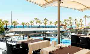 Utsikt från en uteservering mot en poolområde med parasoller och palmer samt havet i bakgrunden. (AI-genererad text)