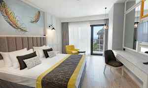 Ett hotellrum med en dubbelsäng, gul fåtölj, skrivbord och spegel, samt balkong med havsutsikt. (AI-genererad text)