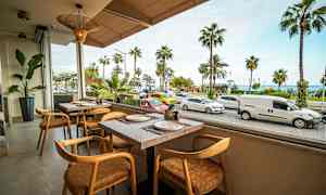 Bilden visar en restaurang med utsikt över en gata med parkerade och förbipasserande bilar samt palmer i bakgrunden. (AI-genererad text)