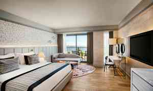 Ett hotellrum med en stor säng, sittgrupp och en balkong mot havet. (AI-genererad text)