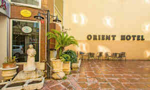 En byggnad med skylten "Orient Hotel" på en ljus vägg, med en liten uteplats dekorerad med stolar, bord, blomkrukor och en staty. (AI-genererad text)