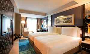 Ett hotellrum med två enkelsängar bredvid varandra, en fåtölj vid ett stort fönster, och ett skrivbord i hörnet. Väggarna är dekorerade med tavlor. (AI-genererad text)