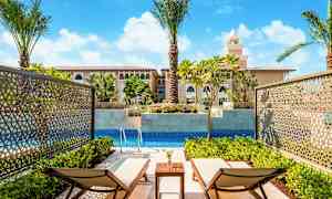 Två solstolar vid en privat pool, omgivna av gröna växter och med utsikt mot en byggnad med medelhavsinspirerade arkitekturelement. (AI-genererad text)