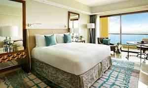 Ett modernt hotellrum med en stor säng, vita och turkosa sängkläder, och en balkong med utsikt över havet. (AI-genererad text)