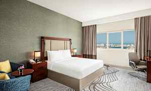 Ett hotellrum med en dubbelsäng, nattduksbord, fåtölj och fönster med utsikt över en stad. (AI-genererad text)