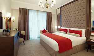 Ett elegant inrett hotellrum med en stor säng klädd i vita och röda sängkläder. Rummet har en stoppad huvudgavel, en kristallkrona och stora fönster med gardiner. (AI-genererad text)