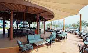 En utomhusbar med sittgrupper och tak i trä, omgiven av palmer och stadssiluetten i bakgrunden. (AI-genererad text)