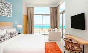 Ett modernt hotellrum med en stor säng, en soffa, skrivbord och en TV. Utsikten från rummet visar en strand och havet. (AI-genererad text)