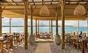 En strandrestaurang med träbord och stolar, utsikt mot havet och en stad i bakgrunden. (AI-genererad text)