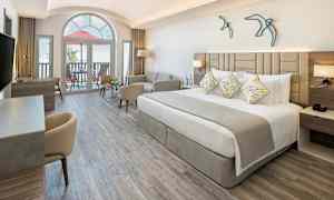 Ett modernt hotellrum med en bred säng, soffa och skrivbord. Stort fönster och balkong i bakgrunden. (AI-genererad text)