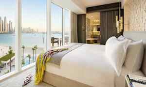 Ett ljust hotellrum med en dubbelsäng, ett öppet badrum och stora fönster som visar en utsikt över havet. (AI-genererad text)