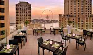 Terrass med dukade bord och stolar, omgiven av höghus med utsikt över ett pariserhjul och havet vid solnedgång. (AI-genererad text)