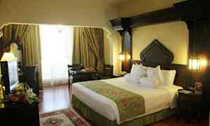 Ett hotellrum med en stor säng, möbler i mörkt trä och fönster täckta med eleganta gardiner. (AI-genererad text)