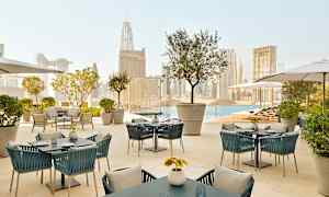 En utomhusterrass med bord och stolar med utsikt över en pool och stadssilhuetten i bakgrunden. (AI-genererad text)