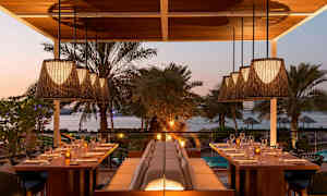 En utomhusrestaurang med utsikt över havet, omgiven av växter och palmer. Bord är dukade med glas och tallrikar, och taklampor hänger ovanför. (AI-genererad text)