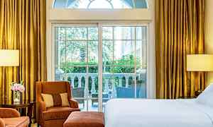 Ett hotellrum med stora fönster som vetter mot en terrass med utsikt över grönska. Rummet har en säng, fåtöljer och guldfärgade gardiner. (AI-genererad text)