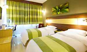 Ett hotellrum med två enkelsängar, dold belysning vid sänggavlarna, en skrivbordsyta och gröna gardiner som kompletterar rummets inredning. (AI-genererad text)