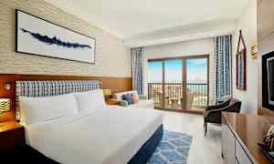 Ett hotellrum med en dubbelsäng, soffa och balkong som vetter mot havet. (AI-genererad text)