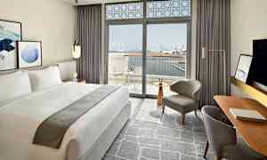 Ett modernt inrett hotellrum med en stor dubbelsäng och fönster som öppnar upp mot en balkong med utsikt över en stadssilhuett och vatten. (AI-genererad text)