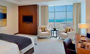 Ett modernt hotellrum med en säng, fåtöljer, ett bord och en arbetsyta. Stora fönster visar utsikten över stad och hav. (AI-genererad text)