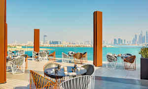 Uteservering med bord och stolar framför en vacker utsikt av stadens skyline och havet. (AI-genererad text)