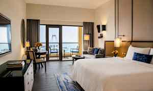 Ett hotellrum med en stor säng, sittgrupp och en matplats. Stora fönster leder ut till en balkong med utsikt över havet och ett stort pariserhjul. (AI-genererad text)