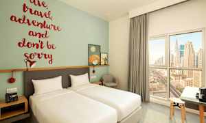 Ett hotellrum med två enkelsängar, en väggdekoration som innehåller ett inspirerande citat, och ett fönster med utsikt över stadens silhuett. (AI-genererad text)