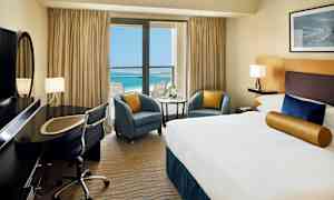 Hotellrum med en stor säng, skrivbord och utsikt över ett blått hav genom en balkong med glasdörrar. (AI-genererad text)