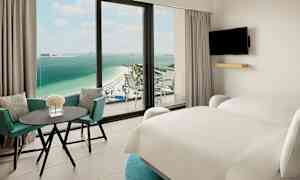 Hotellrum med två sängar, bord och stolar samt utsikt mot en strand och hav. (AI-genererad text)