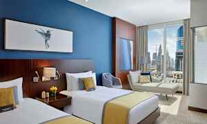 Ett hotellrum med två enkelsängar, en blå vägg med tavlor, en soffa, en fåtölj och ett stort fönster med utsikt över en stad. (AI-genererad text)