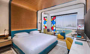 Ett hotellrum med en stor säng, färgglada glasmålningar och utsikt över strand och stad. (AI-genererad text)