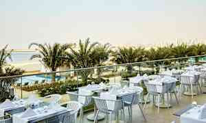 En utomhusrestaurang med dukade bord, med utsikt över palmer, en strand och en stadssiluett i bakgrunden. (AI-genererad text)