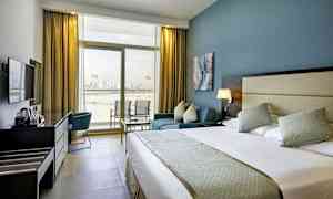 Ett hotellrum med en stor säng, en sittgrupp och en arbetsyta med en vattenkokare. Rummet har ett stort fönster som vetter mot en utsikt över en stad vid vattnet. (AI-genererad text)