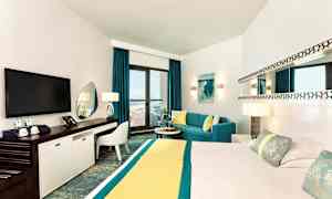 Ett modernt hotellrum med en stor säng, TV, arbetsyta och en sittgrupp vid fönster mot en balkong med havsutsikt. (AI-genererad text)