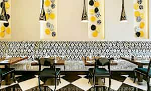 Ett restauranginteriör med svartvita golvplattor, mönstrade väggpaneler, och en dekorativ sittbänk med svarta stolar runt borden. Färggranna väggdekorationer syns på väggen. (AI-genererad text)