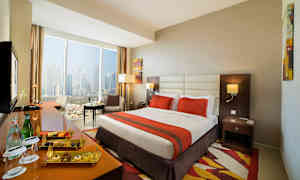 Hotellrum med stor säng, orange överkast, fönster med utsikt över staden och sittgrupp. (AI-genererad text)