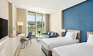 Ett hotellrum med två enkelsängar, en blå vägg, sittgrupp med bord och soffa samt ett stort fönster som visar utsikten. (AI-genererad text)