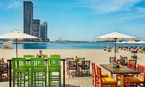 Stolar och bord på en strand med utsikt över havet och stadssiluett i fjärran. (AI-genererad text)