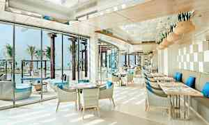 En ljus och modern restaurang med blåa kuddar på stolar och utsikt över en pool och palmer. (AI-genererad text)
