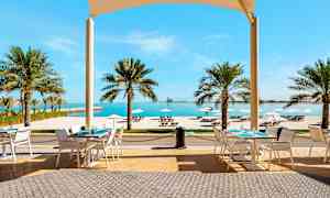 Uteservering med bord, stolar och vy över tropiska palmer, vit sandstrand och hav. (AI-genererad text)