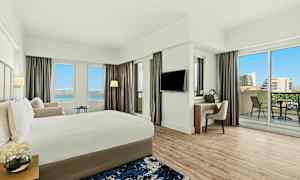 Ett modernt hotellrum med en stor säng, ett skrivbord, balkong och utsikt över en blå himmel och byggnader. (AI-genererad text)