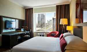 Bild på ett modernt hotellrum med en säng i förgrunden, ett skrivbord med en TV och fönster med utsikt över en stad. (AI-genererad text)