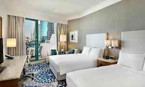 Ett hotellrum med två dubbelsängar, ett skrivbord med stol och en balkong med utsikt över moderna byggnader. (AI-genererad text)