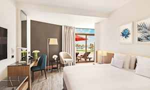 Ett hotellrum med moderna möbler, stor säng, marmorerat skrivbord och en terrass med solstolar och utsikt över palmer. (AI-genererad text)