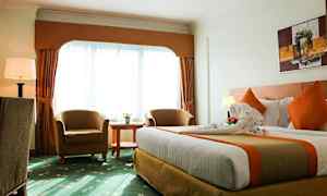 Ett hotellrum med en stor säng, två fåtöljer, ett runt bord och en stor fönsteryta med gardiner. (AI-genererad text)