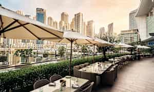 En uteservering med parasoller och bord vid en hamn med höghus i bakgrunden. (AI-genererad text)