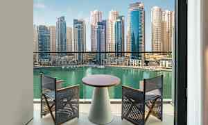 Utsikt från en balkong mot en turkos vattenyta, kantad av moderna höghus. På balkongen finns ett bord och två stolar. (AI-genererad text)