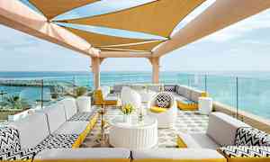 En uteplats med sittgrupper och ett tak skuggat av tyg framför en havsutsikt. (AI-genererad text)