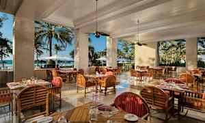 Restaurang med trämöbler och utsikt mot stranden genom stora öppningar. (AI-genererad text)